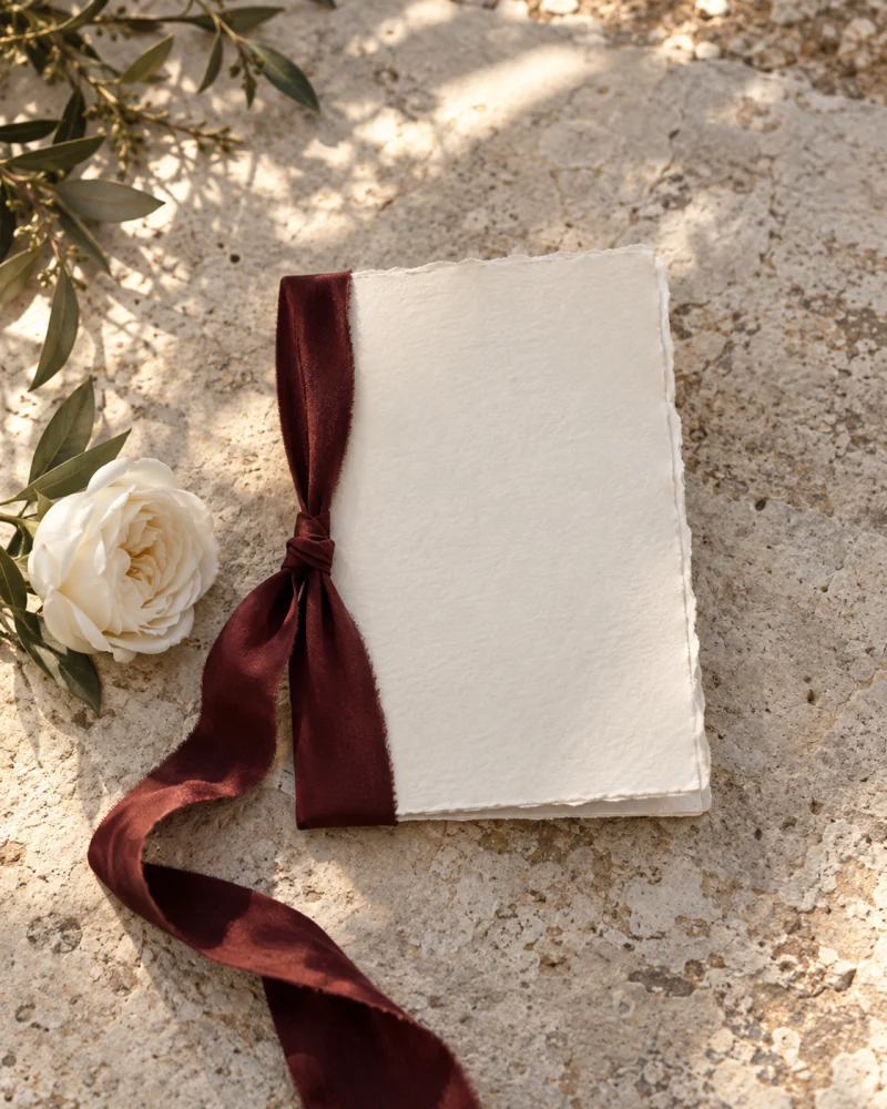 An ivory vow booklet with a burgundy silk ribbon and a white rose